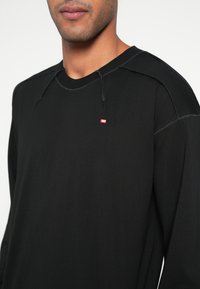 Juoda ilgomis rankovėmis marškinėliai su apvalia iškirpte, matomomis siūlėmis ir mažu raudonu Diesel logotipu ant krūtinės. Minkšta, lengva medžiaga.