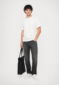 Grå jeans, hvit t-skjorte og hvite sneakers. Personen holder en svart tote bag. Enkelt bakgrunn, uformell stil, minimalistisk design.