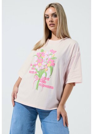 Kobieta w różowej, za dużej koszulce z kolorową grafiką bukietu i napisem "Wszystko, czego potrzebujesz, to miłość i kwiaty", w zestawieniu z niebieskimi dżinsami.
