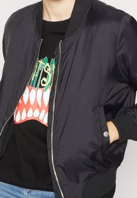 Veste bomber noire avec poignets et col côtelés, fermeture éclair à l'avant, poches latérales, et matériau lisse et brillant. En dessous, un t-shirt graphique.