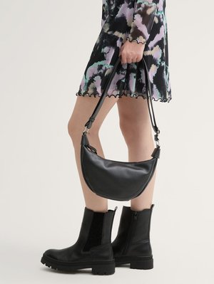 Femme portant des bottines noires et une robe mi-longue à motifs abstraits, tenant un sac à main en cuir noir par sa bandoulière.