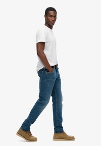 Camiseta blanca de manga corta, vaqueros de mezclilla azul y zapatos casuales de color marrón claro. El modelo está de pie de manera casual con las manos en los bolsillos.