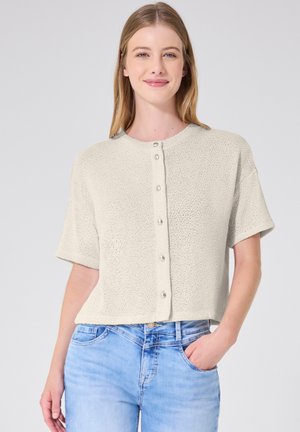 Ung kvinde med langt blondt hår iført en kortærmet cremefarvet strikket skjorte med knapper og lyseblå jeans med den ene hånd i lommen.