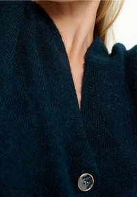 Mörkgrön, texturerad stickad cardigan med djup v-halsringning och en stor svart knapp som stängning. Mjukt material, avslappnad design.