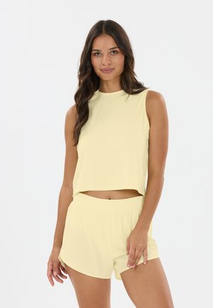 Jeune femme aux longs cheveux foncés portant un crop top sans manches jaune clair et un short assorti, debout devant un fond blanc uni.