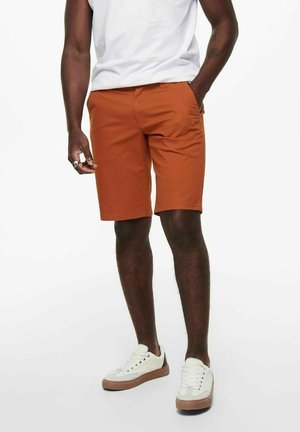 Uomo che indossa pantaloncini color ruggine fino al ginocchio, sneakers bianche con suola in gomma e una maglietta bianca, in piedi con una mano in tasca.