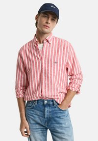 Rot- und weiß gestreiftes Langarmhemd mit Button-Down-Kragen, über einem weißen T-Shirt getragen, kombiniert mit hellblauen Jeans. Marineblaues Cap auf dem Kopf.