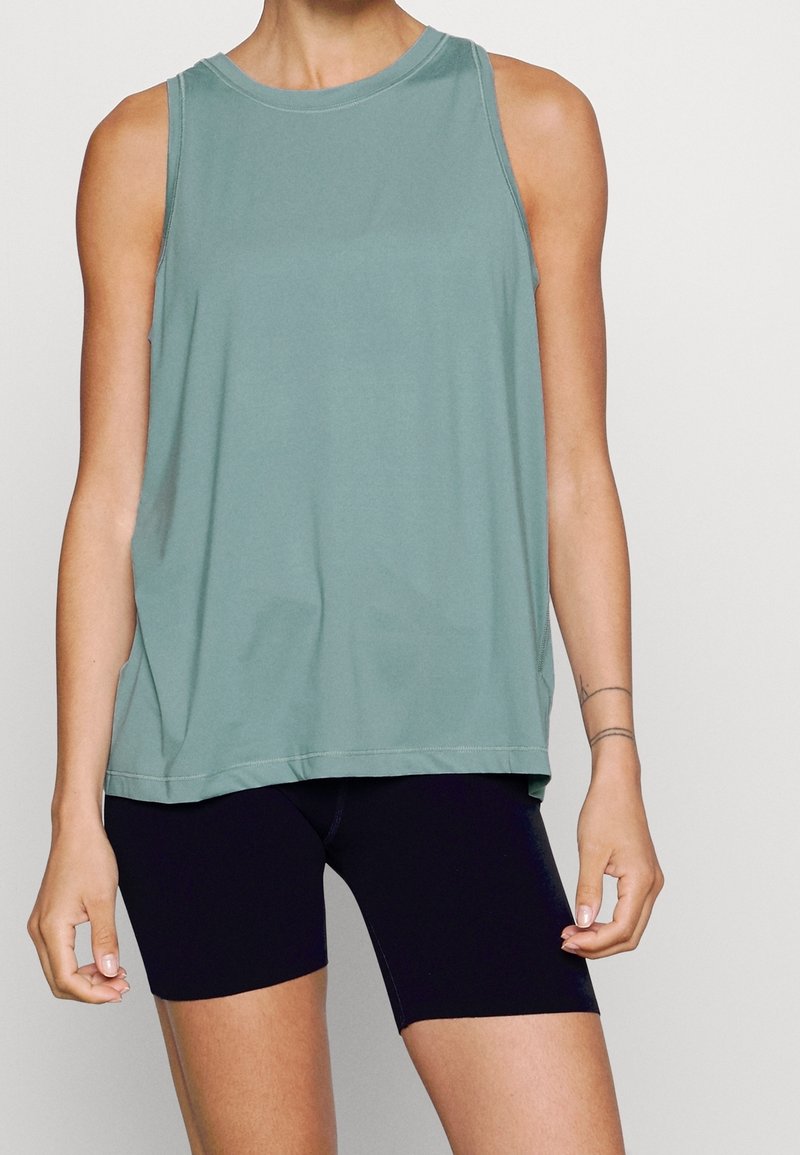 Femme portant un haut sans manches vert clair et un short noir ajusté devant un fond uni.