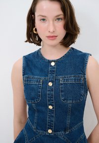 Robe en denim avec un corsage ajusté, sans manches et deux poches avant. Comprend des fermetures à boutons en ton doré et une jupe légèrement évasée.