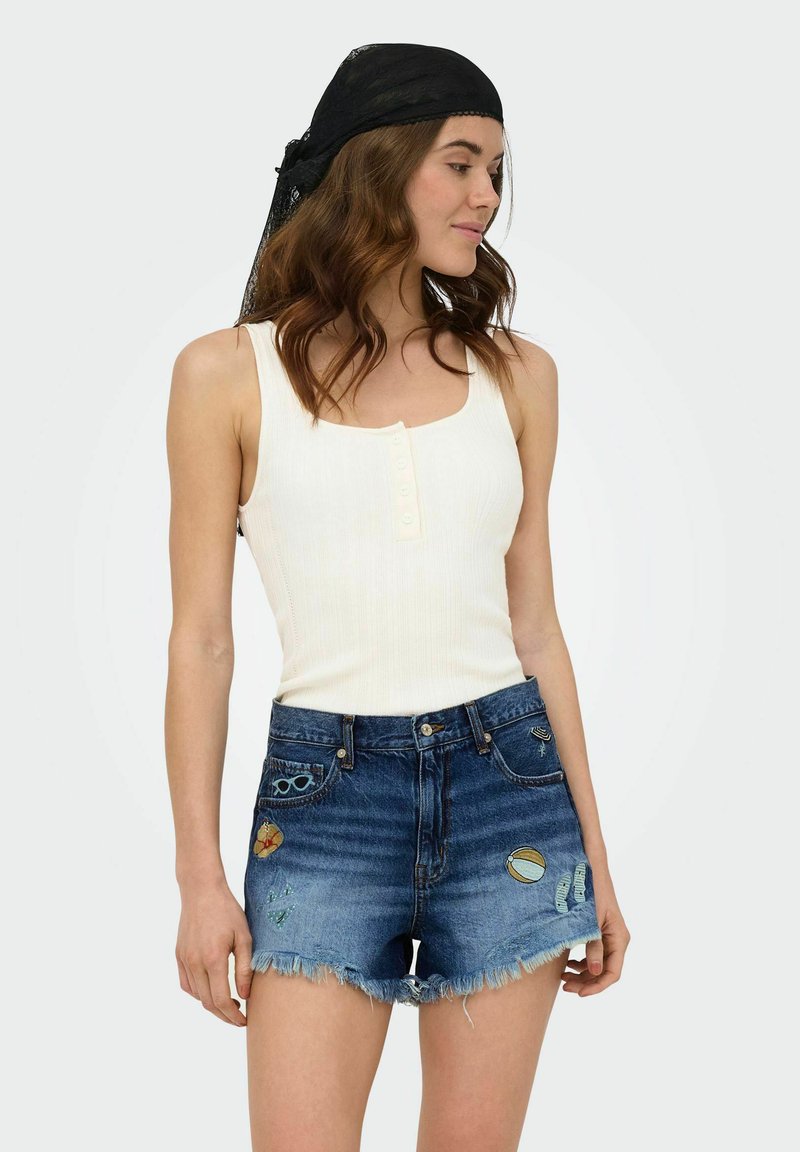 Femme portant un haut blanc sans manches, un short en jean bleu avec des patchs et un foulard noir sur la tête, debout devant un fond uni.