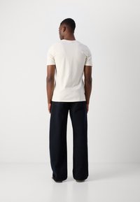 Tricou crem cu mânecă scurtă, realizat din bumbac, asociat cu pantaloni largi negri, văzut din spate pe un fundal gri deschis.