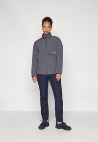 Giacca grigia con zip e colletto alto, abbinata a pantaloni blu navy e scarpe da trekking nere. Tessuto liscio con design minimalista.