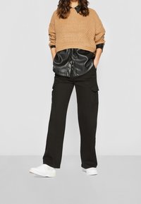 Pull en maille beige par-dessus une chemise en cuir noire, associé à un pantalon cargo noir ample et des baskets blanches. Tenue décontractée en superposition avec des textures variées.