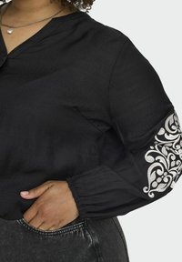 Bluză neagră cu decolteu în V și mâneci lungi, având broderie decorativă argintie pe mâna stângă și manșete plisate.