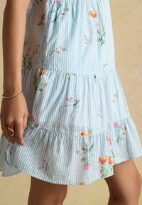 Seitenansicht einer Person, die ein blau-weiß gestreiftes Kleid mit Blumenmustern, ein goldenes Armband und einen Ring am linken Hand trägt.