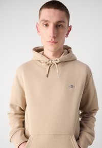 Beige hooded sweatshirt with drawstring, featuring a small embroidered logo on the chest and ribbed cuffs and hem. Smooth cotton material.