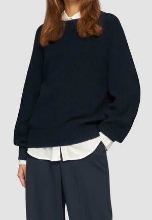 Kobieta ubrana w granatowy sweter z teksturowanego dzianiny na białej koszuli zapinanej na guziki oraz granatowe spodnie o szerokich nogawkach, ręce w kieszeniach, na szarym tle.