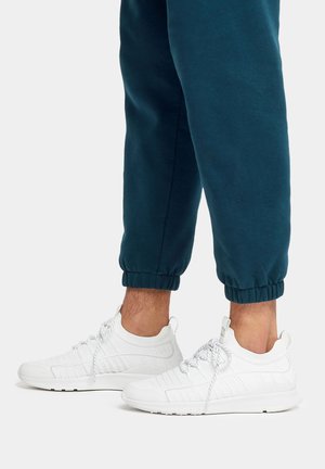 Weiße Sportschuhe mit strukturierten Überzügen und gemusterten Seiten, ausgestattet mit reflektierenden Schnürsenkeln und einer abgerundeten Spitze, kombiniert mit dunklen Jogginghosen.