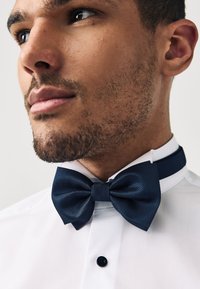 Papillon blu navy con texture e fascia abbinata, indossato su una camicia bianca con bottone nero, che mostra un design formale.