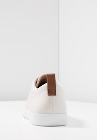 Valkoinen nahkakengät litteällä pohjalla, jossa on ruskea nahkavetotappi kannassa ja minimaalisia ompelutietoja.