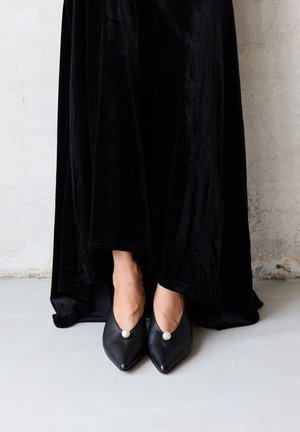 Nogi w czarnej aksamitnej spódnicy, noszące czarne szpiczaste szpilki z pojedynczą dużą perłową dekoracją, stojące na szarej podłodze na tle jasnej, teksturowanej ściany.