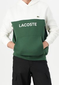 Felpa con cappuccio Lacoste verde e bianca con un grande logo bianco, tasca a marsupio e cappuccio con coulisse. Realizzata in un tessuto morbido e strutturato.