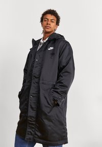 Manteau rembourré noir avec capuche, doté d'une fermeture éclair sur le devant, d'une fermeture à boutons-pression et de poches latérales. Logo sur la poitrine gauche, texture lisse.