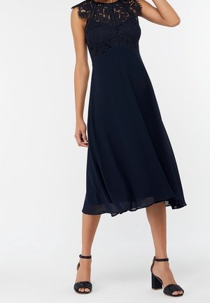 Granatowa midi sukienka bez rękawów z koronkową górą i zwiewną spódnicą, zestawiona z czarnymi czółenkami na słupku z paskiem wokół kostki.