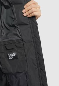 Giacca nera imbottita, raffigurante un primo piano di una mano che tiene il tessuto, con un'etichetta logo visibile e una texture liscia.