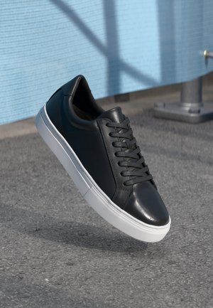 Svart lær sneaker med rund tå, hvit gummisåle og flate skolisser. Glatt tekstur og minimalistisk design.