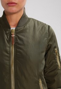Béžová bomber bunda olivové barvy se zipem na přední straně, žebrovaným límcem a dvěma bočními kapsami. Disponuje zlatým kováním a hladkým saténovým povrchem.
