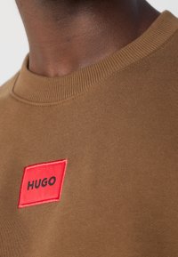 Gros plan sur un t-shirt marron à col rond avec un patch rectangulaire rouge portant l'inscription « HUGO » sur la poitrine, porté par une personne à la peau foncée.
