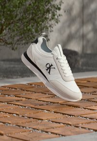 Λευκό αθλητικό παπούτσι Calvin Klein με μαύρο λογότυπο, λευκά κορδόνια και μαύρη λεπτομέρεια στη φτέρνα, αιωρούμενο πάνω από ξύλινο δάπεδο έξω.