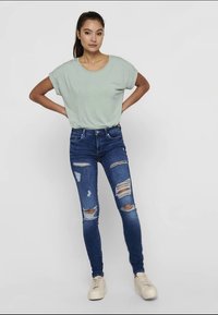 T-shirt oversized verde claro, jeans skinny azuis desfiados com vários rasgos e ténis bege, exibidos contra um fundo neutro.