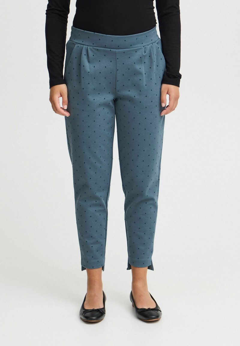 Türaunfarbene, gemusterte Hose mit schwarzen Punkten, hoher Taille und schmal zulaufenden Beinen, kombiniert mit schwarzen flachen Schuhen.