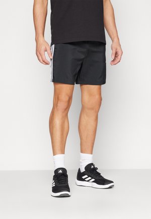 Man draagt zwarte sportshorts met witte zijkstrepen, zwart-witte Adidas sneakers en witte sportsokken, staand op een witte vloer.