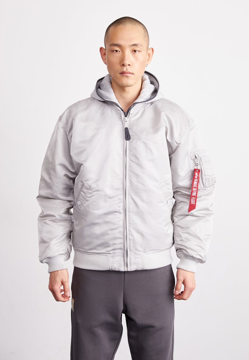 Veste bomber gris clair avec fermeture éclair à l'avant, poignets côtelés et capuche, avec une étiquette rouge sur la manche. Texture de tissu lisse et brillant.
