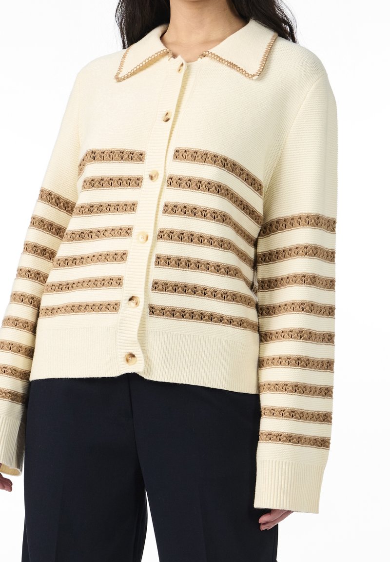 Roomkleurige cardigan met een kraag, voorzien van horizontale bruine strepen en decoratieve stiksels, gesloten met lichte knopen.
