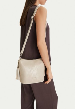 Donna che indossa un abito scuro senza maniche, con una borsa a tracolla beige testurizzata con dettaglio a frange sulla spalla.