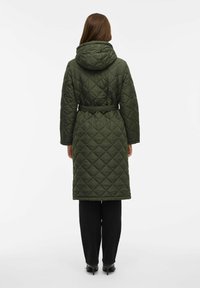 O femeie îmbrăcată într-un palton lung, matlasat, de culoare verde măslinie, cu glugă și curea, pantaloni negri și pantofi cu toc negri, stând cu spatele.