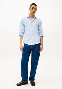 Lichtblauw button-up overhemd met lange mouwen, gecombineerd met blauwe denim jeans en donkerbruine instappers. Rechte voorkant en een relaxte pasvorm.
