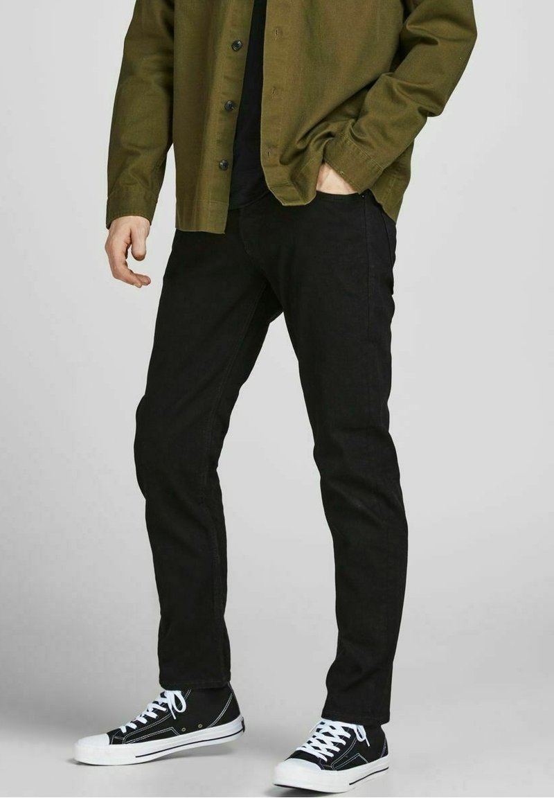 Jeans negros ajustados, de material de algodón, combinados con una camisa de botones corta en verde oliva y zapatillas altas negras con suelas blancas.