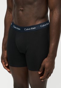 Ανδρικός κορμός που φοράει μαύρα μπόξερ Calvin Klein με ναυτικό μπλε ζωνάκι και λευκό λογότυπο, τα χέρια χαλαρά στο πλάι σε ουδέτερο φόντο.
