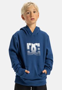 Modrá mikina s kapucňou a prednou klokankou, spevnenými rukávmi a veľkým bielym logom DC na hrudi. Mäkkučký materiál a rebrované manžety.