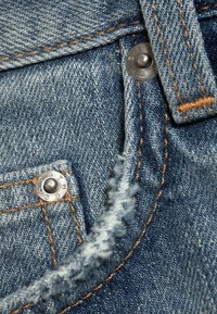 Gros plan d’un jean en denim bleu montrant des rivets métalliques, des surpiqûres orange, une boucle de ceinture et un bord effiloché près de la poche.
