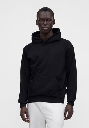 Hombre de pie con sudadera negra con capucha y pantalones blancos sobre un fondo claro y liso.