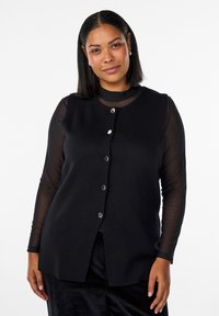 Vrouw met zwart steil haar draagt zwarte mouwloze vest met knopen over een doorschijnende top met lange mouwen, staand tegen een witte achtergrond.