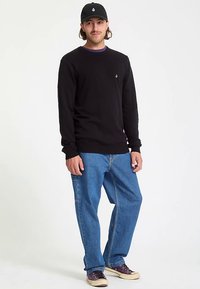 Black long-sleeve shirt with a small logo on the chest, paired with blue denim jeans and black cap. Model has a relaxed stance.