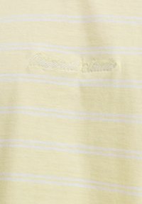 Tissu jaune pâle avec de fines rayures horizontales blanches et un texte brodé blanc pâle indiquant « Wetpaint’s classic. »