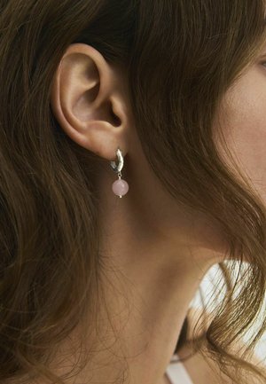 Prim-plan cu urechea unei femei cu păr castaniu ondulat, purtând un cercel argintiu cu un pandantiv rotund roz.